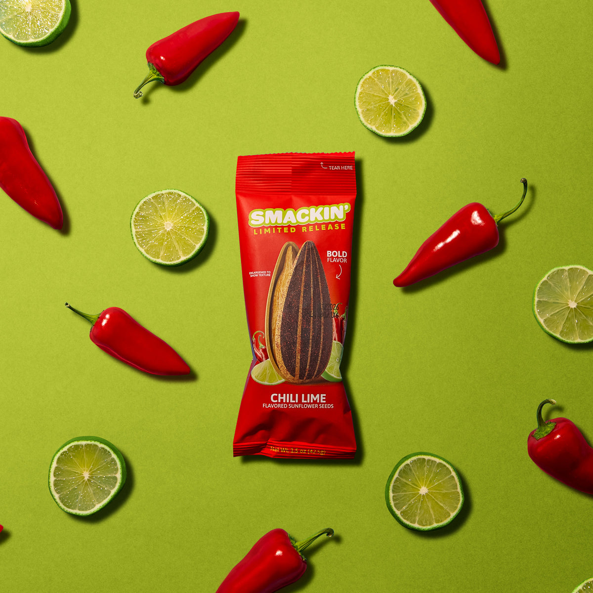 Product photo of a red SMACKIN' Limited Release Chili Lime flavored sunflower seeds bag with an enlarged seed graphic, surrounded by vibrant red chili peppers and fresh green lime slices on a bright lime green background