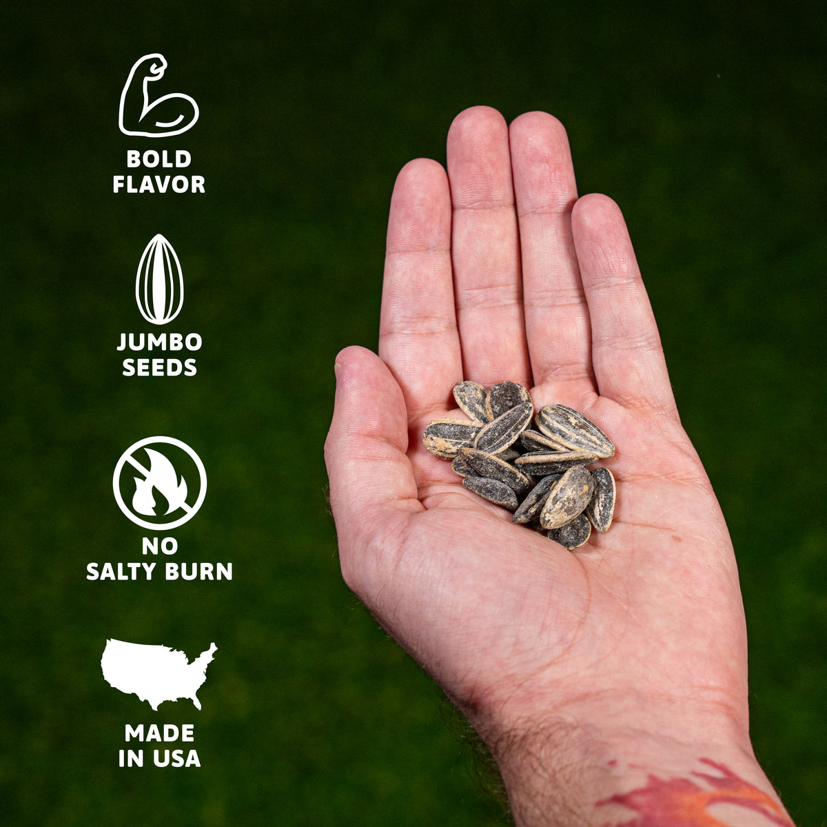 Photograph depicting a person's hand cupping numerous dark roasted sunflower seeds against a textured dark green background, with white textual overlays and icons on the left side reading 'Bold Flavor,' 'Jumbo Seeds,' 'No Salty Burn,' and 'Made in USA,' and a hint of a red and orange design on the wrist