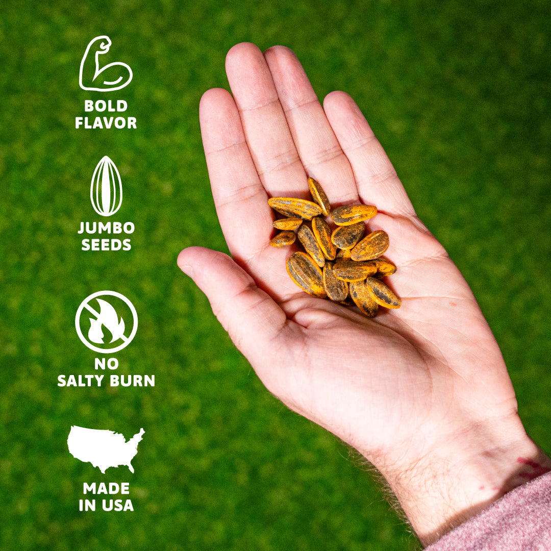 A human hand holds a generous amount of vibrant orange-yellow coated sunflower seeds against a bright green background, accompanied by white icons signifying bold flavor, jumbo seeds, no salty burn, and Made in USA