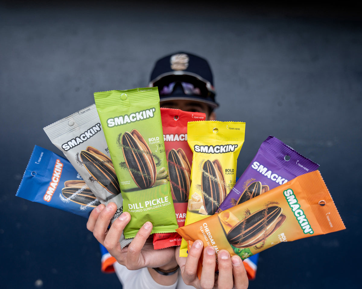 Photograph of a person in a baseball cap and jersey holding seven vibrant bags of Smackin' sunflower seeds, including Dill Pickle, Cheddar Jack, and Cheddar Jalapeno flavors, against a dark blue wall