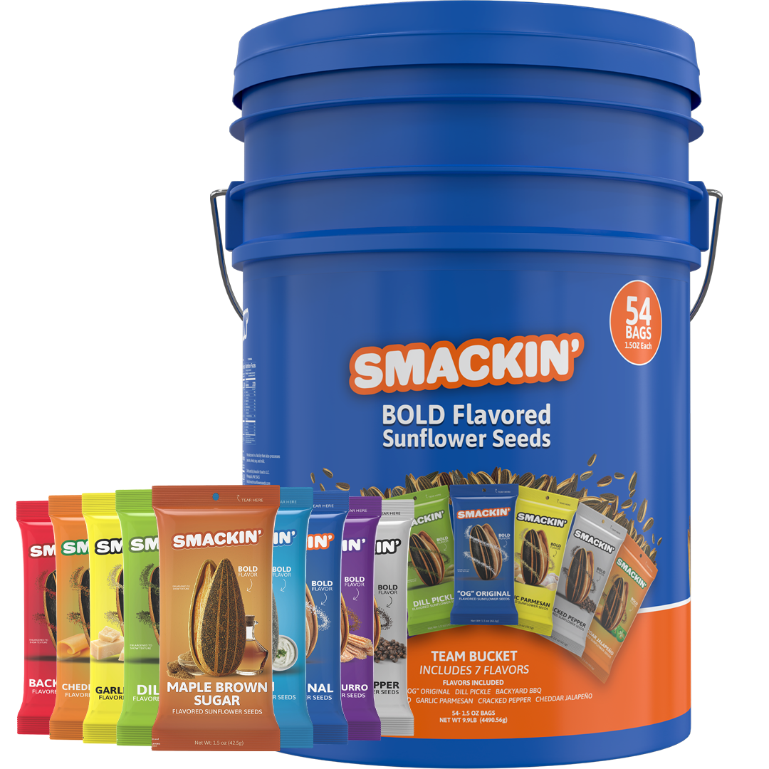 A studio product photo showing a large blue Smackin' Team Bucket containing 54 bold flavored sunflower seed bags, with an assortment of colorful individual packets including Maple Brown Sugar, Dill Pickle, and Cheddar Jalapeno flavors fanned out in front