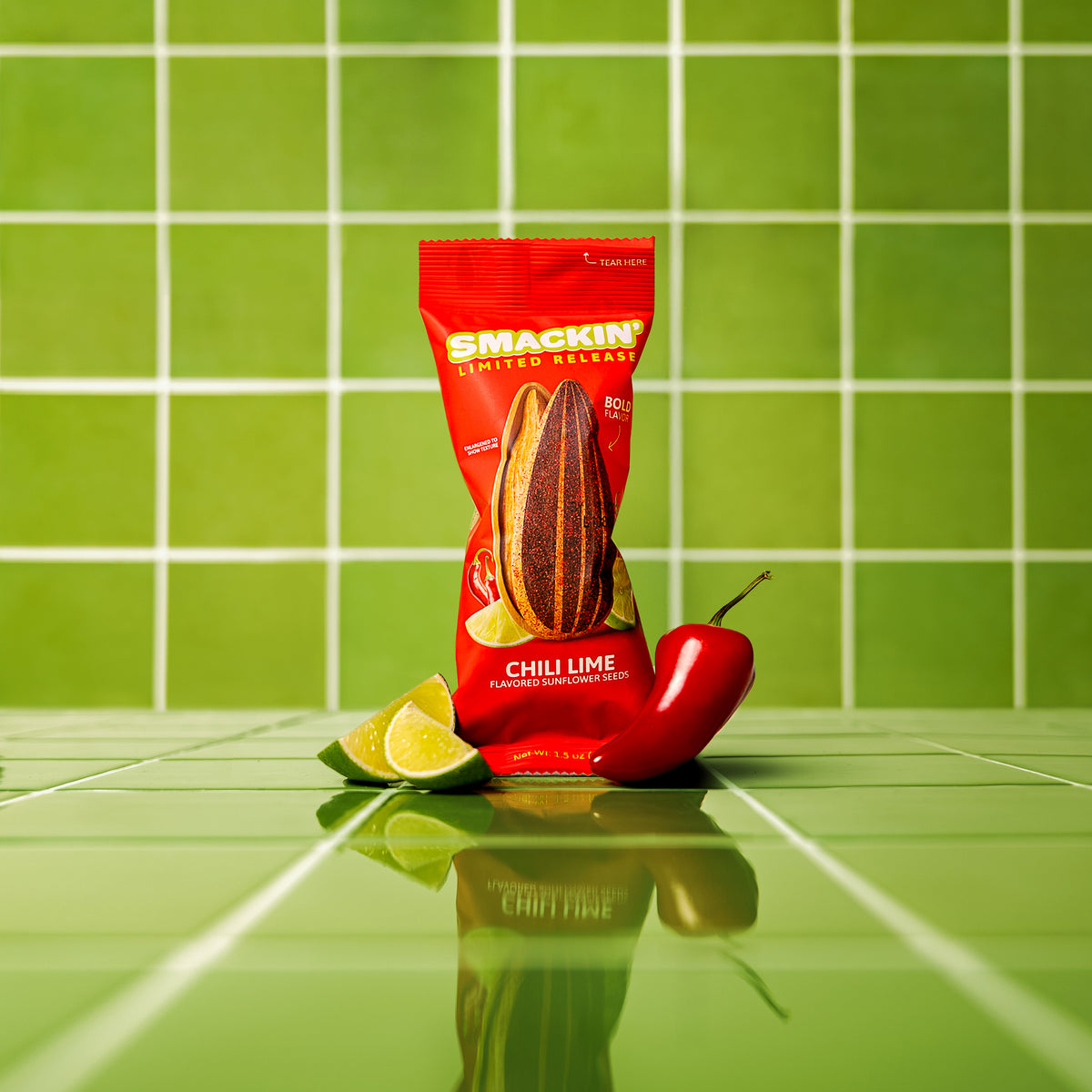 A vibrant studio product photo displays a red Smackin' Limited Release Chili Lime sunflower seeds bag featuring a large illustrative sunflower seed alongside two green lime wedges and a red chili pepper, all resting on a reflective bright green tiled surface with white grout lines