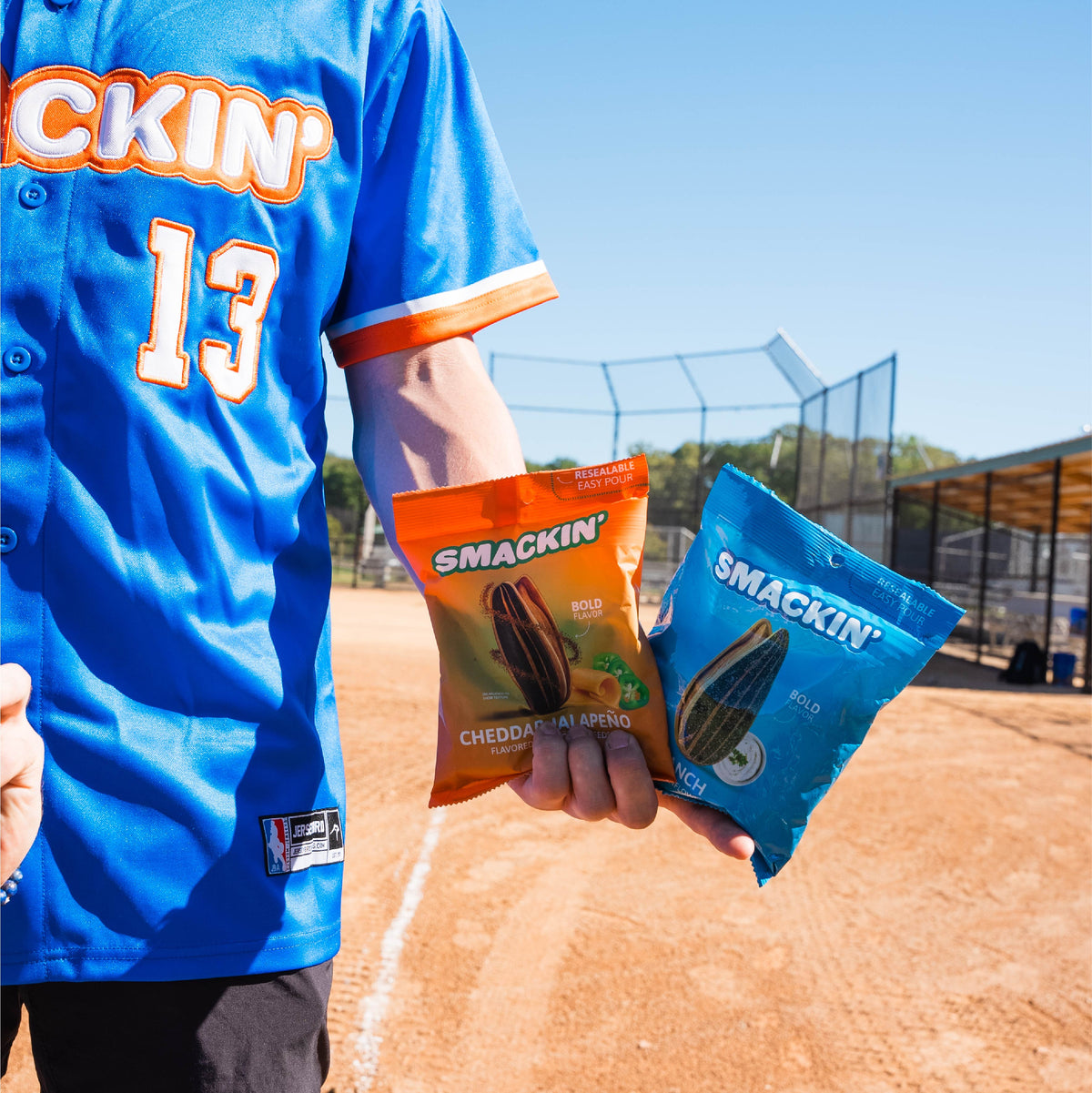 A person in a blue baseball jersey featuring "CKIN' 13" holds an orange Smackin' Cheddar Jalapeño and a blue Smackin' Ranch sunflower seed bag on a red dirt baseball field with a clear blue sky and backstop net visible