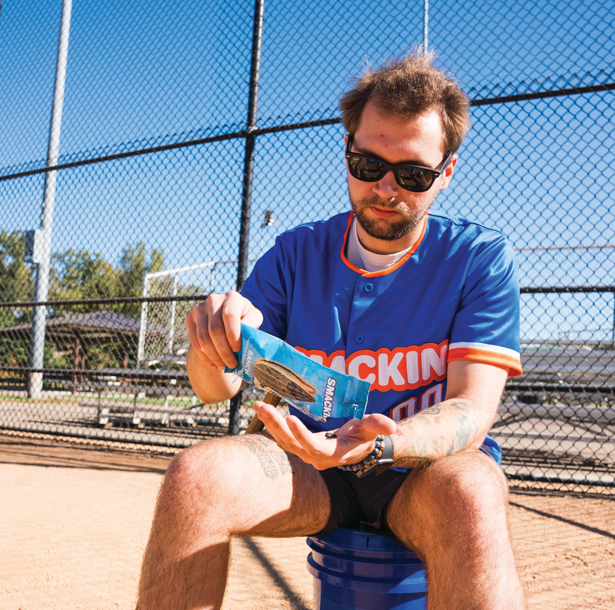 Photograph of a man in a blue baseball jersey with orange trim and sunglasses sitting on a blue bucket on a dirt baseball field, pouring Ranch flavored SMACKIN' sunflower seeds from a blue packet into his hand against a chain-link fence background