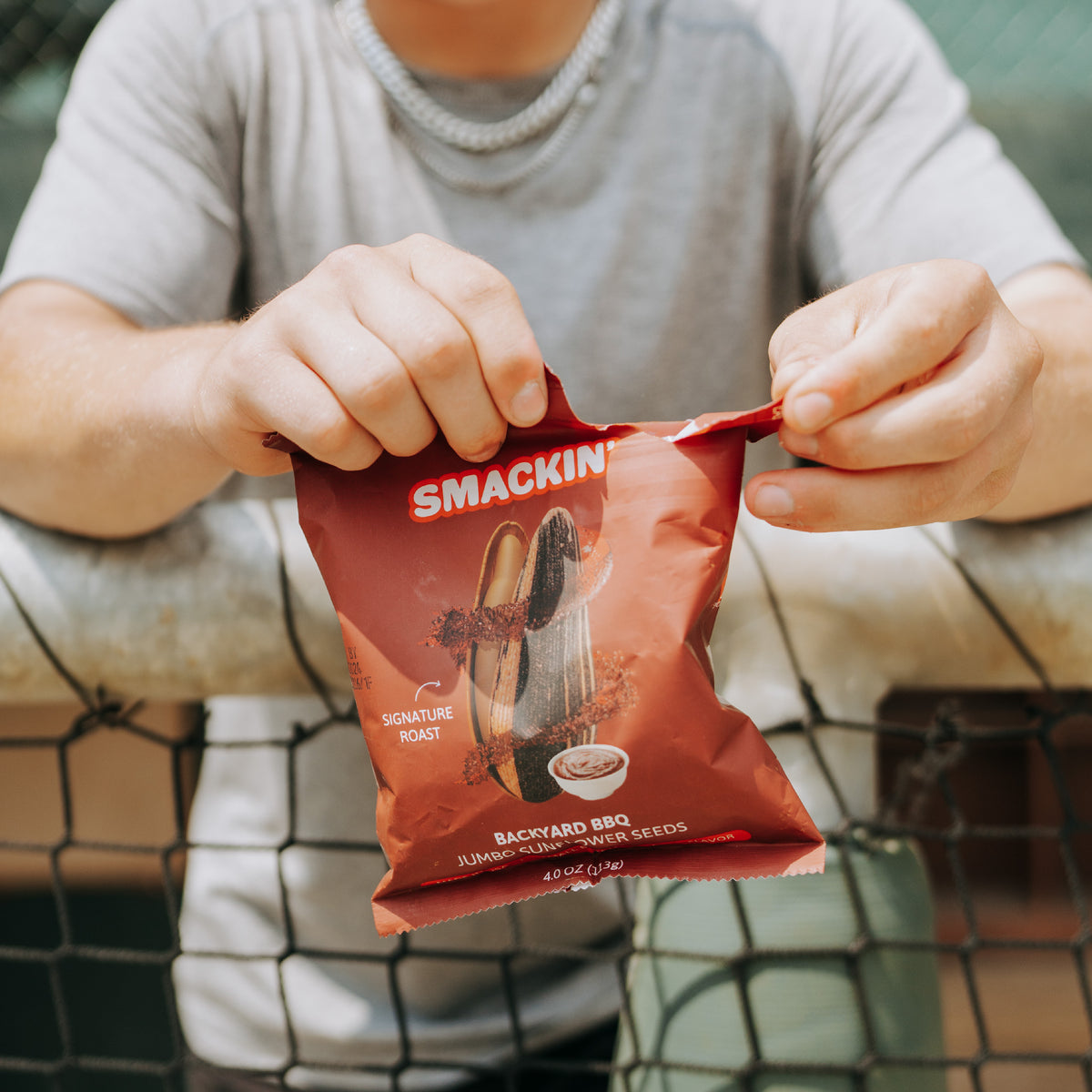A person’s hands in a grey shirt open a red bag of SMACKIN’ Backyard BBQ Jumbo Sunflower Seeds against a blurred chain-link fence at a sports venue