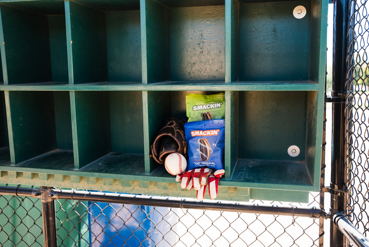 Photograph of a baseball dugout interior featuring a brown baseball glove a white baseball with red stitching red and white batting gloves and vibrant green and blue Smackin' sunflower seed bags stored in dark green cubbies with a chain-link fence in the foreground