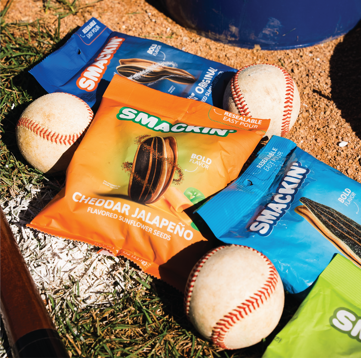 Colorful Smackin' sunflower seed bags, including orange Cheddar Jalapeño and blue Original flavors, scattered on a vibrant baseball field with three worn baseballs, a wooden bat, and a blue dugout container